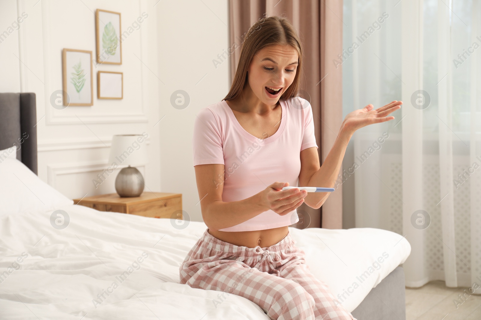 Emotional young woman with pregnancy test on bed at home Photo of Emotional young woman with pregnancy test on bed at home