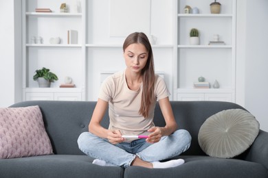Photo of Worried young woman with pregnancy test on sofa at home