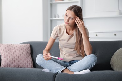Photo of Worried young woman with pregnancy test on sofa at home