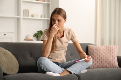Photo of Worried young woman with pregnancy test on sofa at home