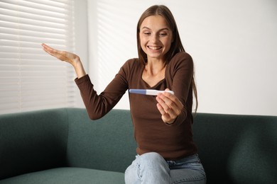 Photo of Happy young woman with pregnancy test on sofa at home
