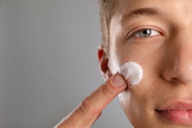 Photo of Handsome young man applying cream onto his face on grey background, closeup. Space for text