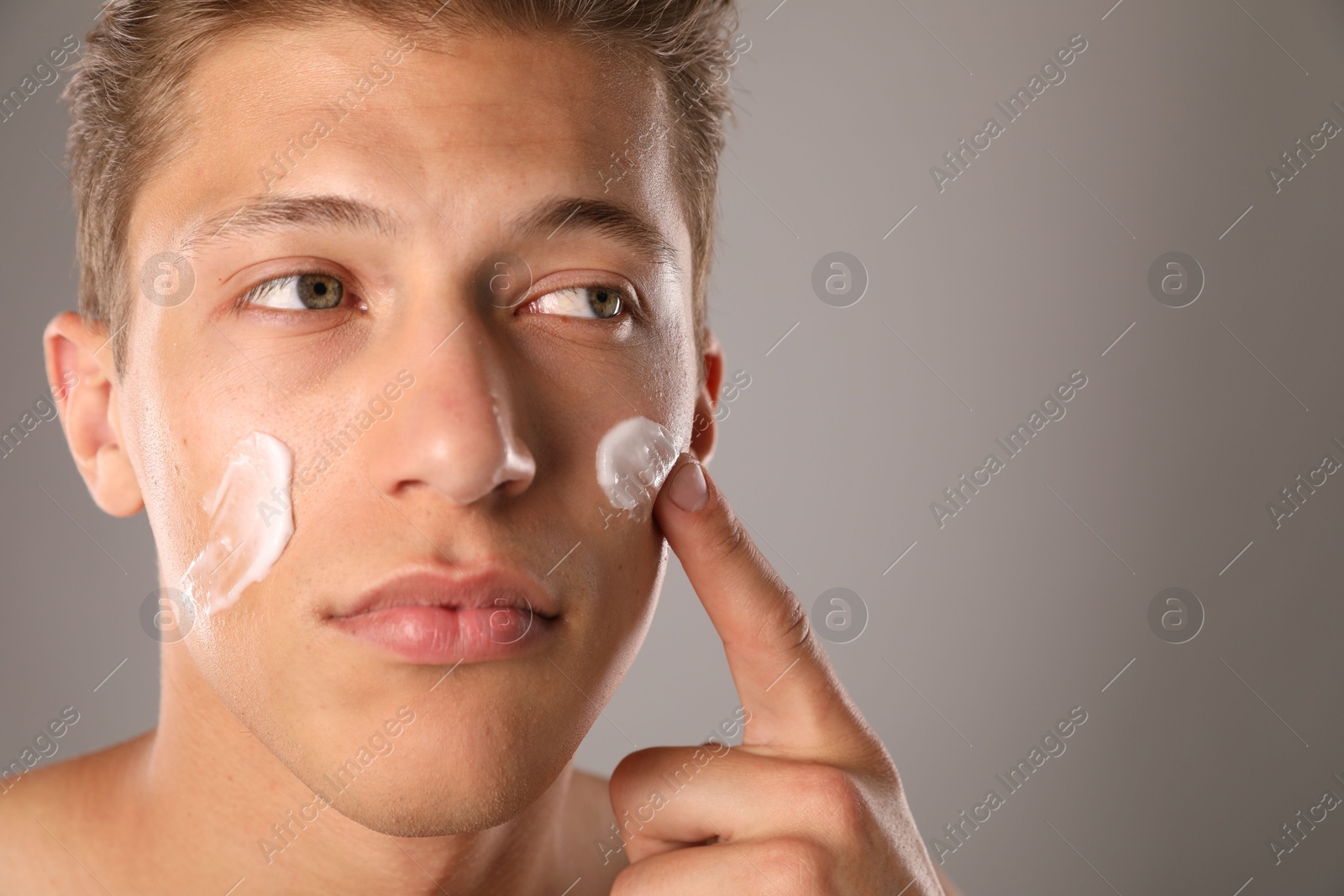 Photo of Handsome young man applying cream onto his face on grey background, closeup. Space for text