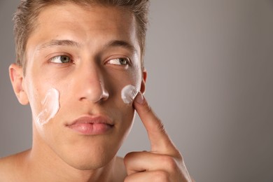 Photo of Handsome young man applying cream onto his face on grey background, closeup. Space for text