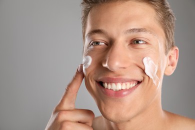 Photo of Handsome young man applying cream onto his face on grey background, closeup