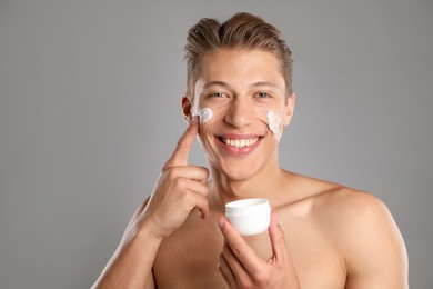 Photo of Handsome young man applying cream onto his face on grey background