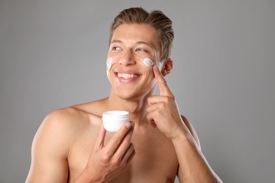 Photo of Handsome young man applying cream onto his face on grey background