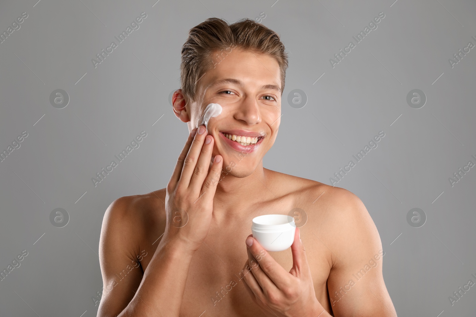Photo of Handsome young man applying cream onto his face on grey background