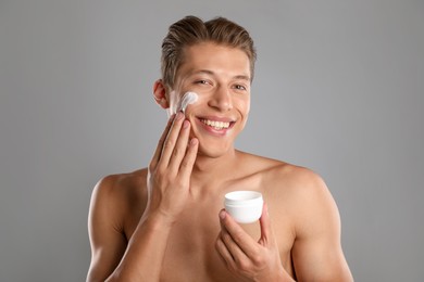 Photo of Handsome young man applying cream onto his face on grey background