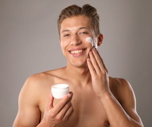 Photo of Handsome young man applying cream onto his face on grey background