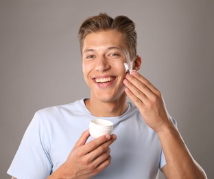 Photo of Handsome young man applying cream onto his face on grey background