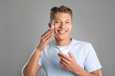 Photo of Handsome young man applying cream onto his face on grey background