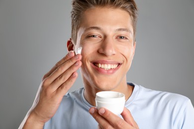 Photo of Handsome young man applying cream onto his face on grey background