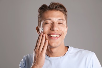 Photo of Handsome young man applying cream onto his face on grey background