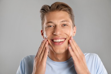 Photo of Handsome young man applying cream onto his face on grey background