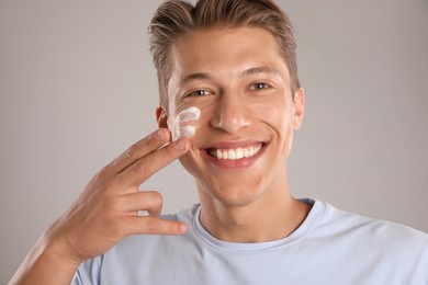 Photo of Handsome young man applying cream onto his face on grey background