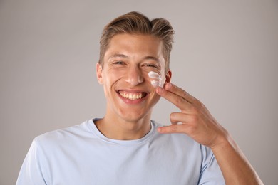 Photo of Handsome young man applying cream onto his face on grey background