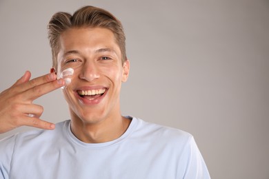 Photo of Handsome young man applying cream onto his face on grey background, space for text