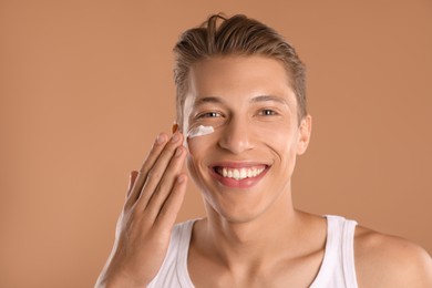 Photo of Handsome young man applying cream onto his face on light brown background