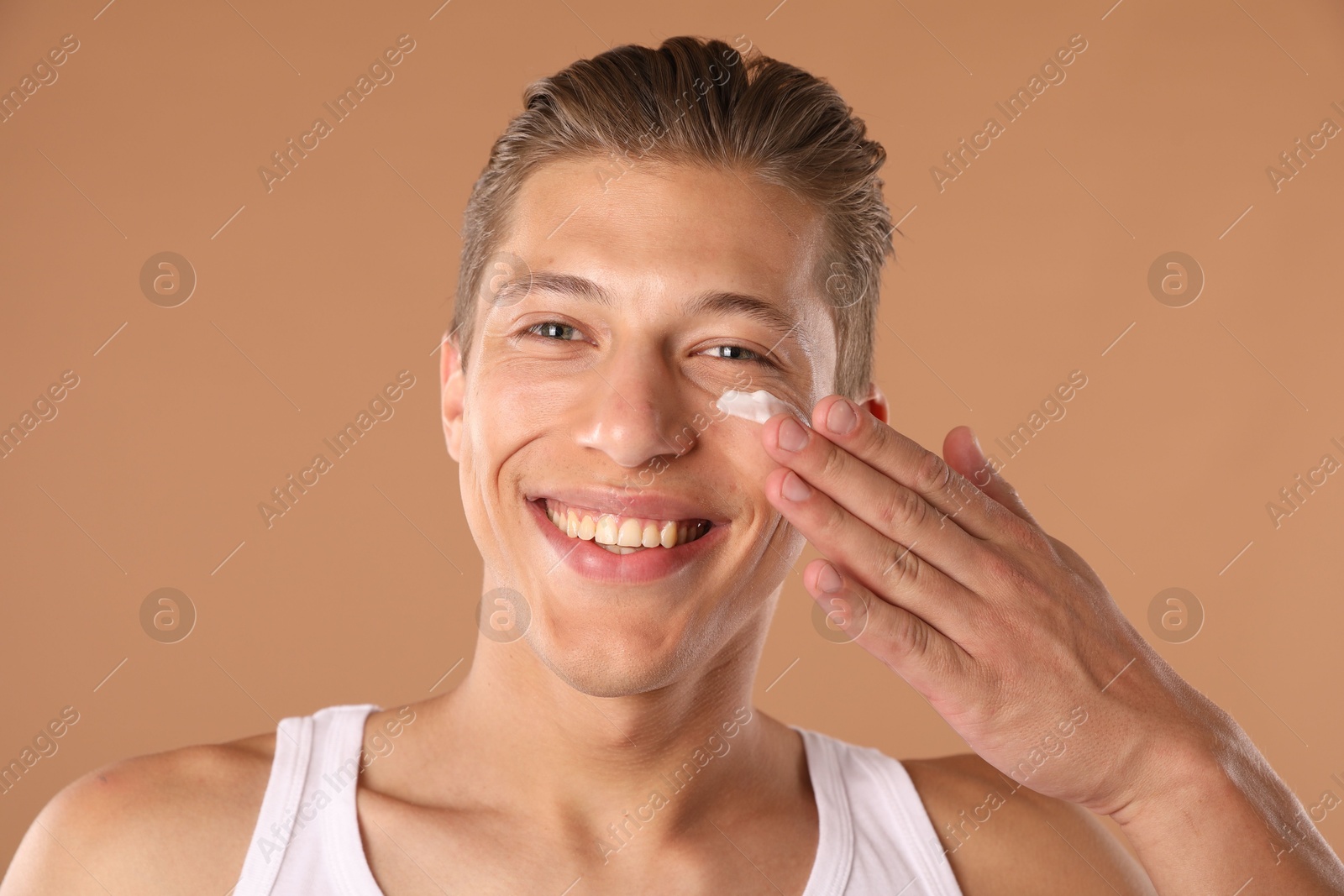 Photo of Handsome young man applying cream onto his face on light brown background