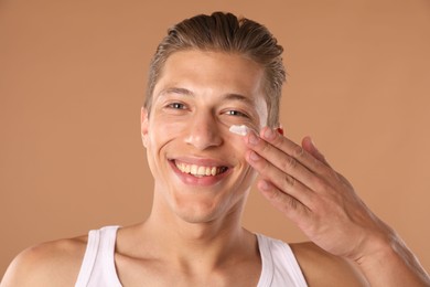 Photo of Handsome young man applying cream onto his face on light brown background