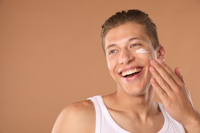 Photo of Handsome young man applying cream onto his face on light brown background, space for text