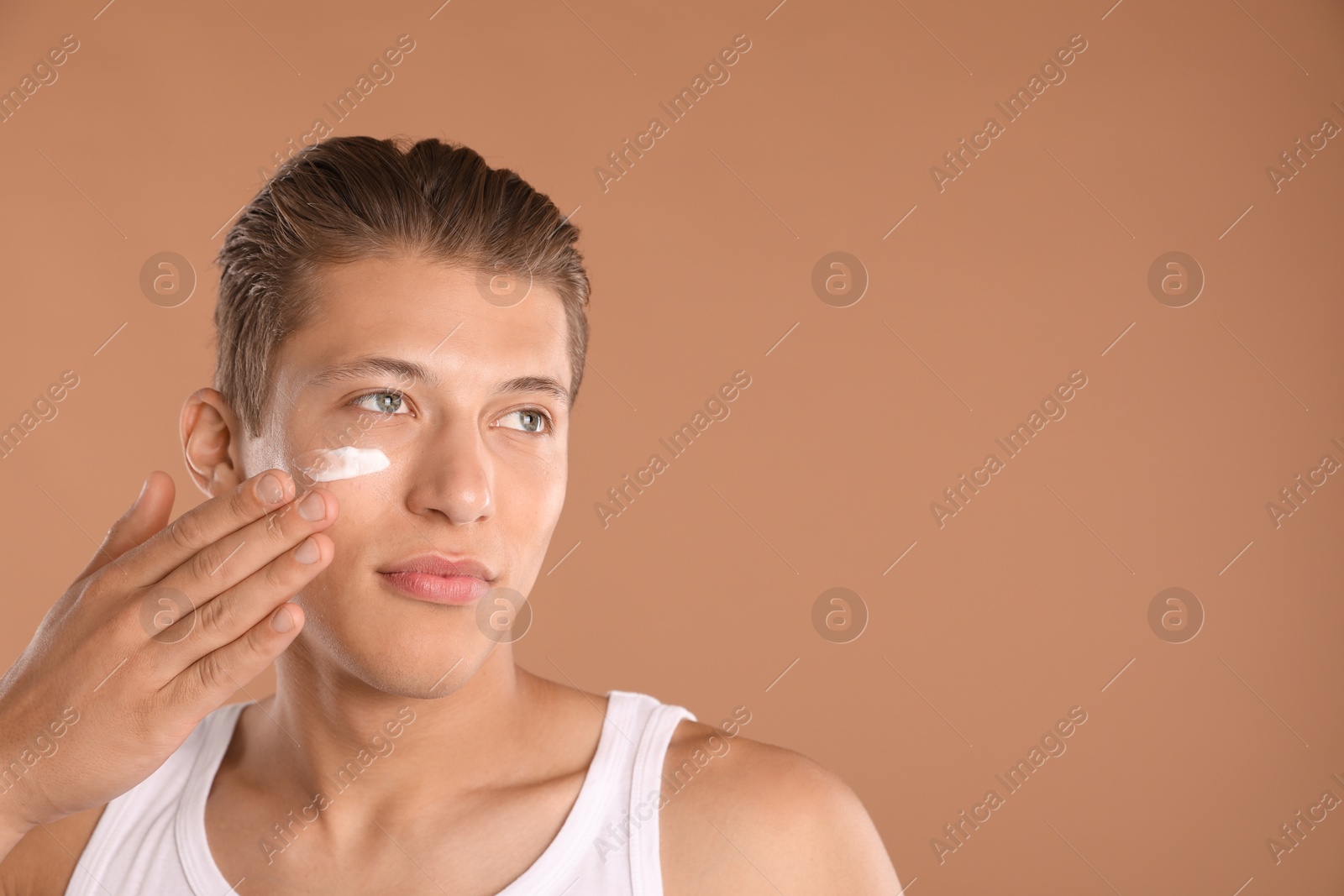 Photo of Handsome young man applying cream onto his face on light brown background, space for text