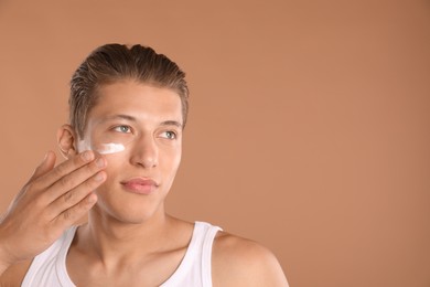 Photo of Handsome young man applying cream onto his face on light brown background, space for text