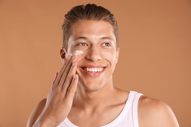 Photo of Handsome young man applying cream onto his face on light brown background