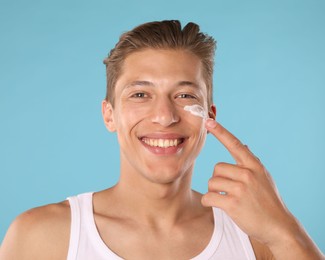 Photo of Handsome young man applying cream onto his face on light blue background