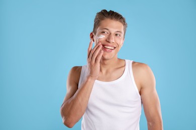 Photo of Handsome young man applying cream onto his face on light blue background
