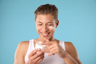 Photo of Handsome young man with jar of face cream on light blue background