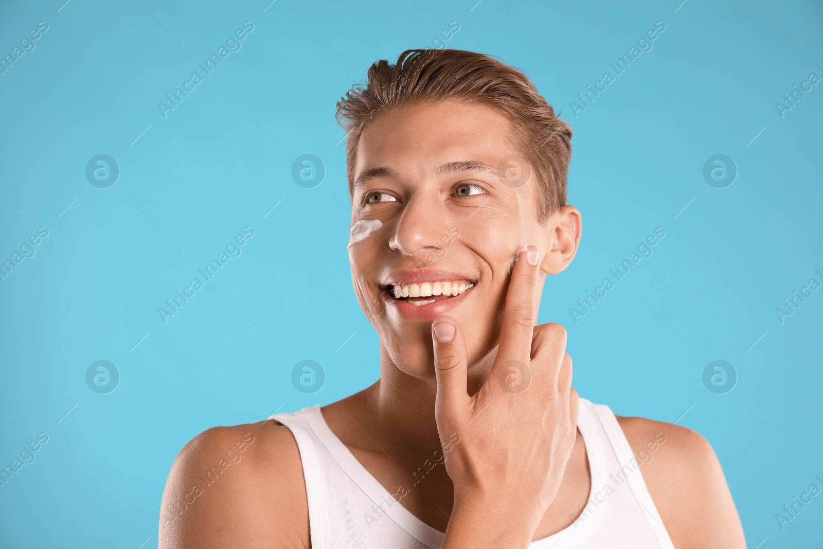 Photo of Handsome young man applying cream onto his face on light blue background