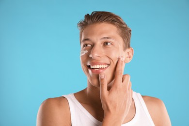 Photo of Handsome young man applying cream onto his face on light blue background