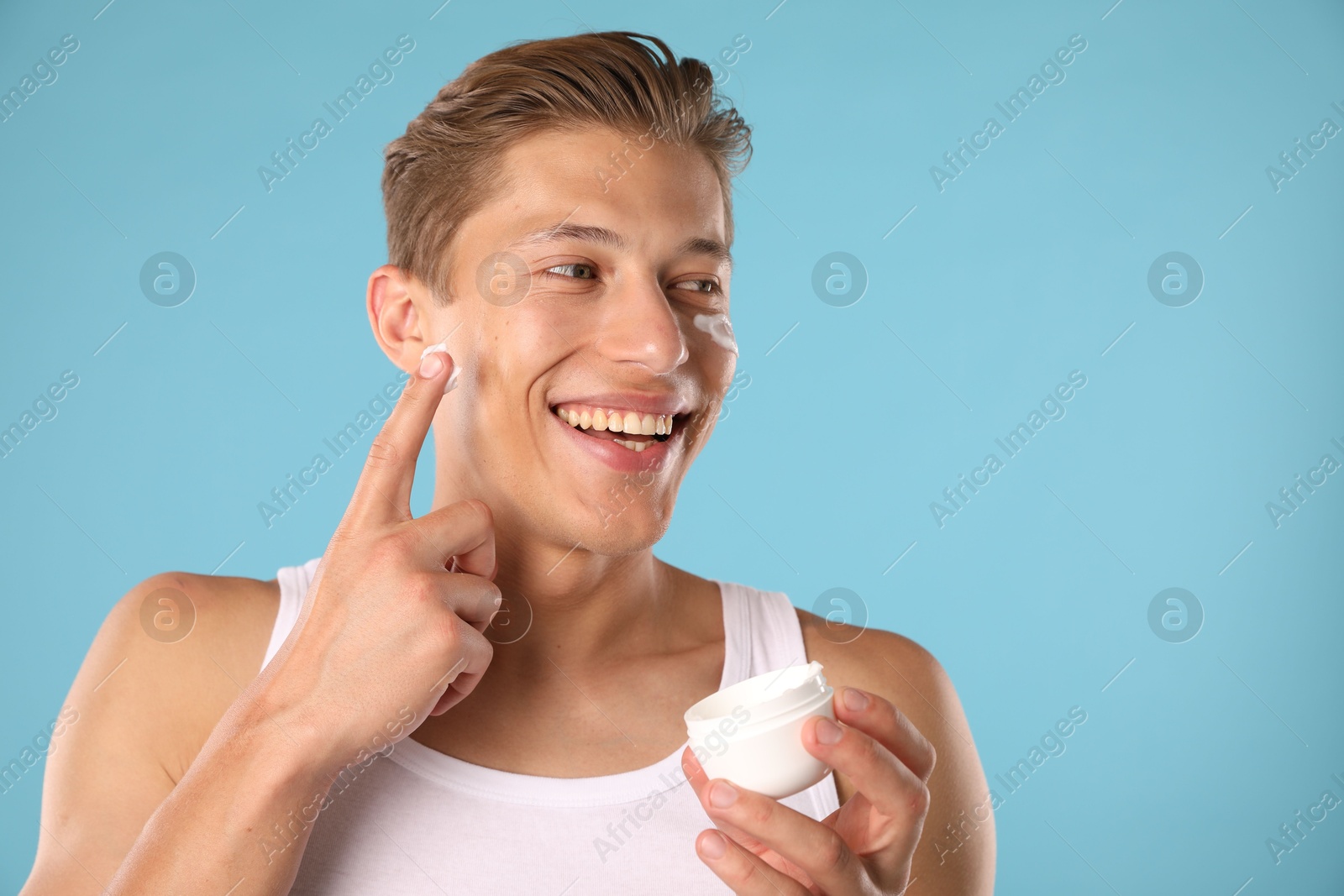 Photo of Handsome young man applying cream onto his face on light blue background