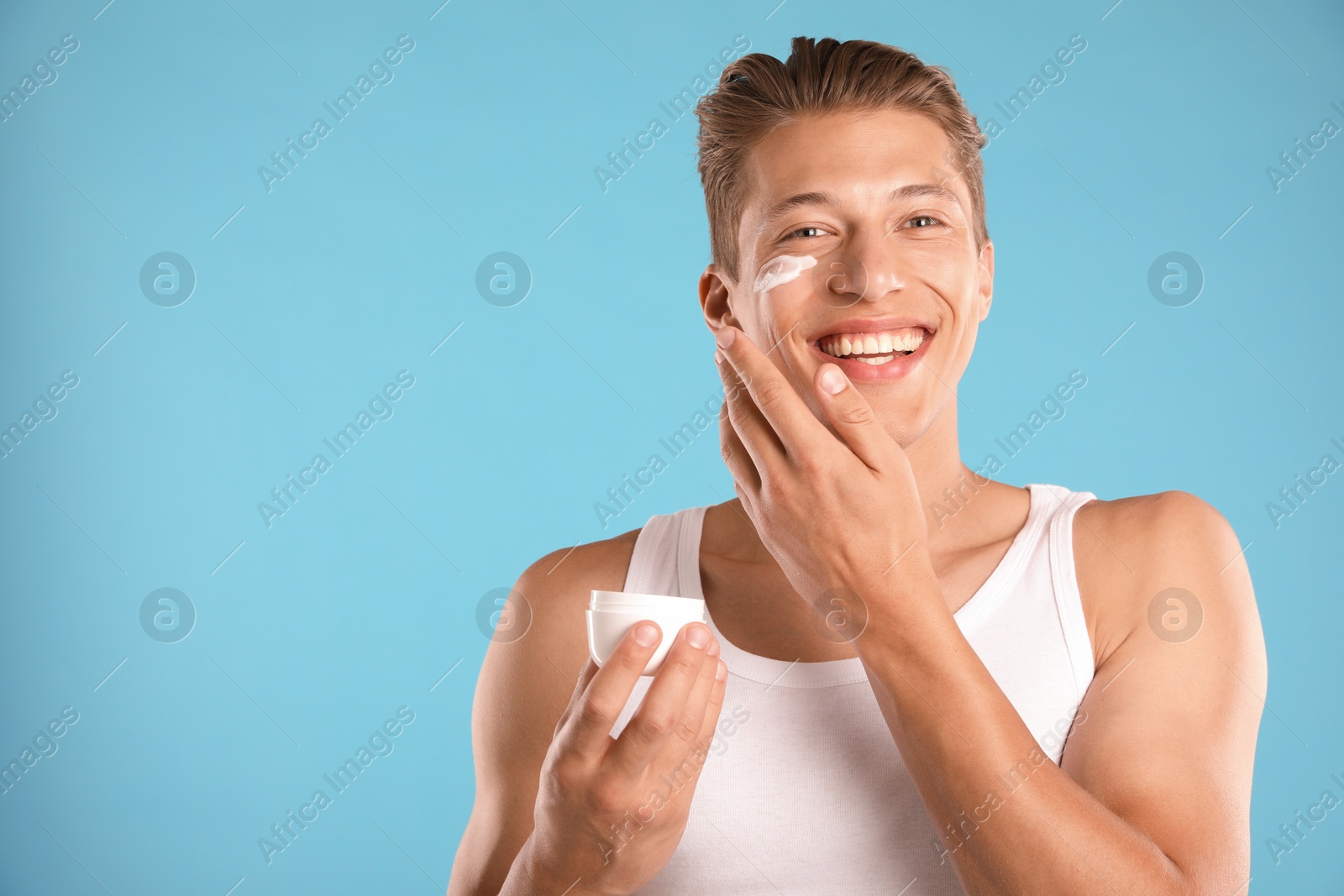 Photo of Handsome young man applying cream onto his face on light blue background. Space for text