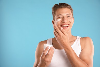 Photo of Handsome young man applying cream onto his face on light blue background. Space for text