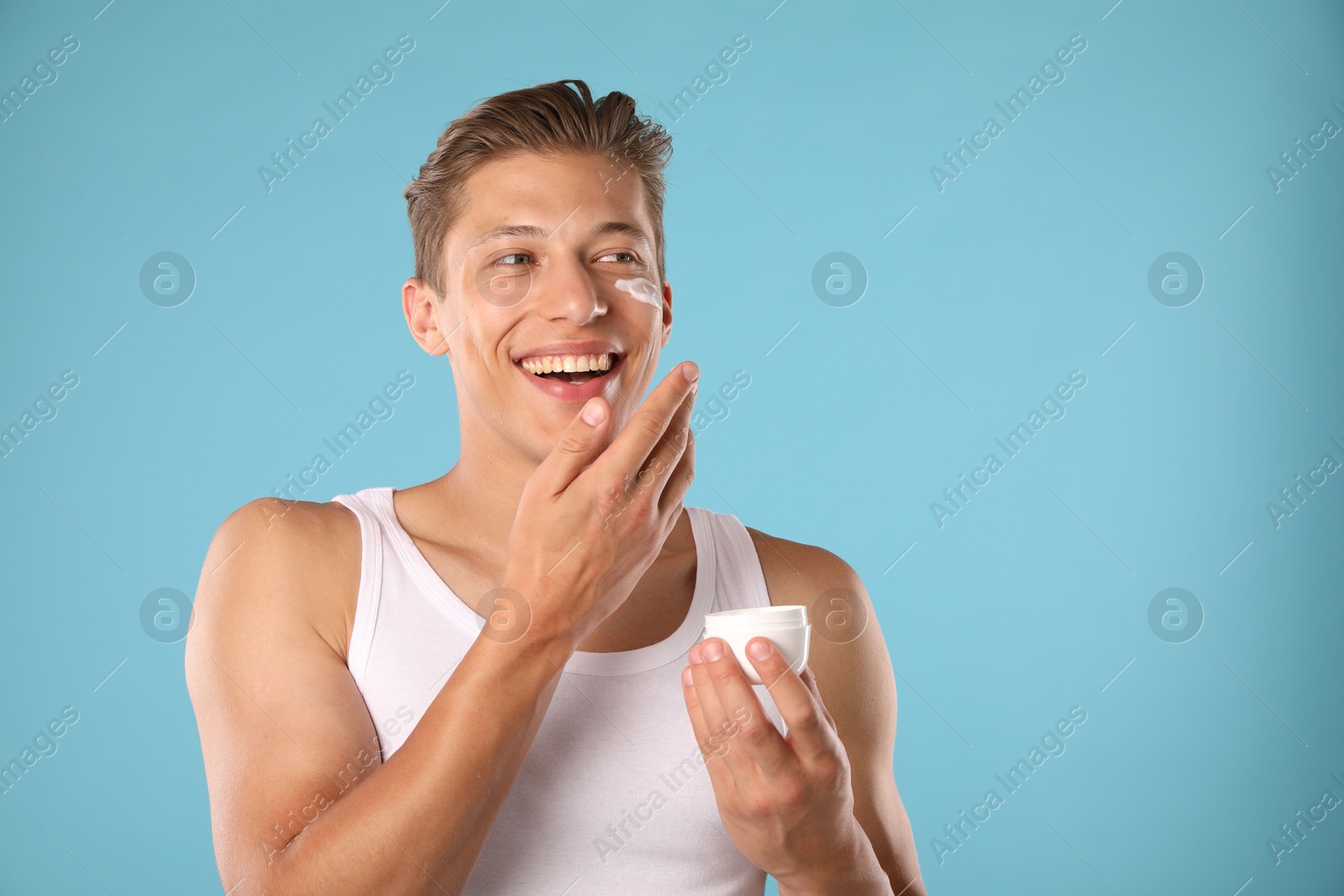 Photo of Handsome young man applying cream onto his face on light blue background. Space for text