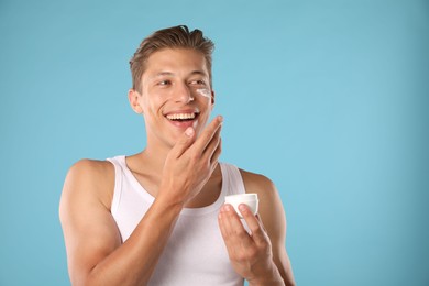 Photo of Handsome young man applying cream onto his face on light blue background. Space for text