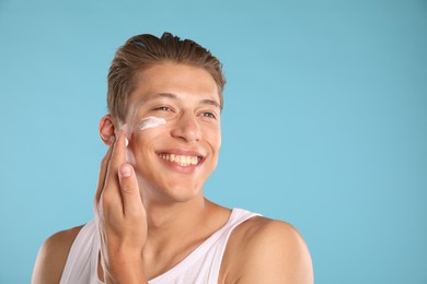 Photo of Handsome young man applying cream onto his face on light blue background, space for text