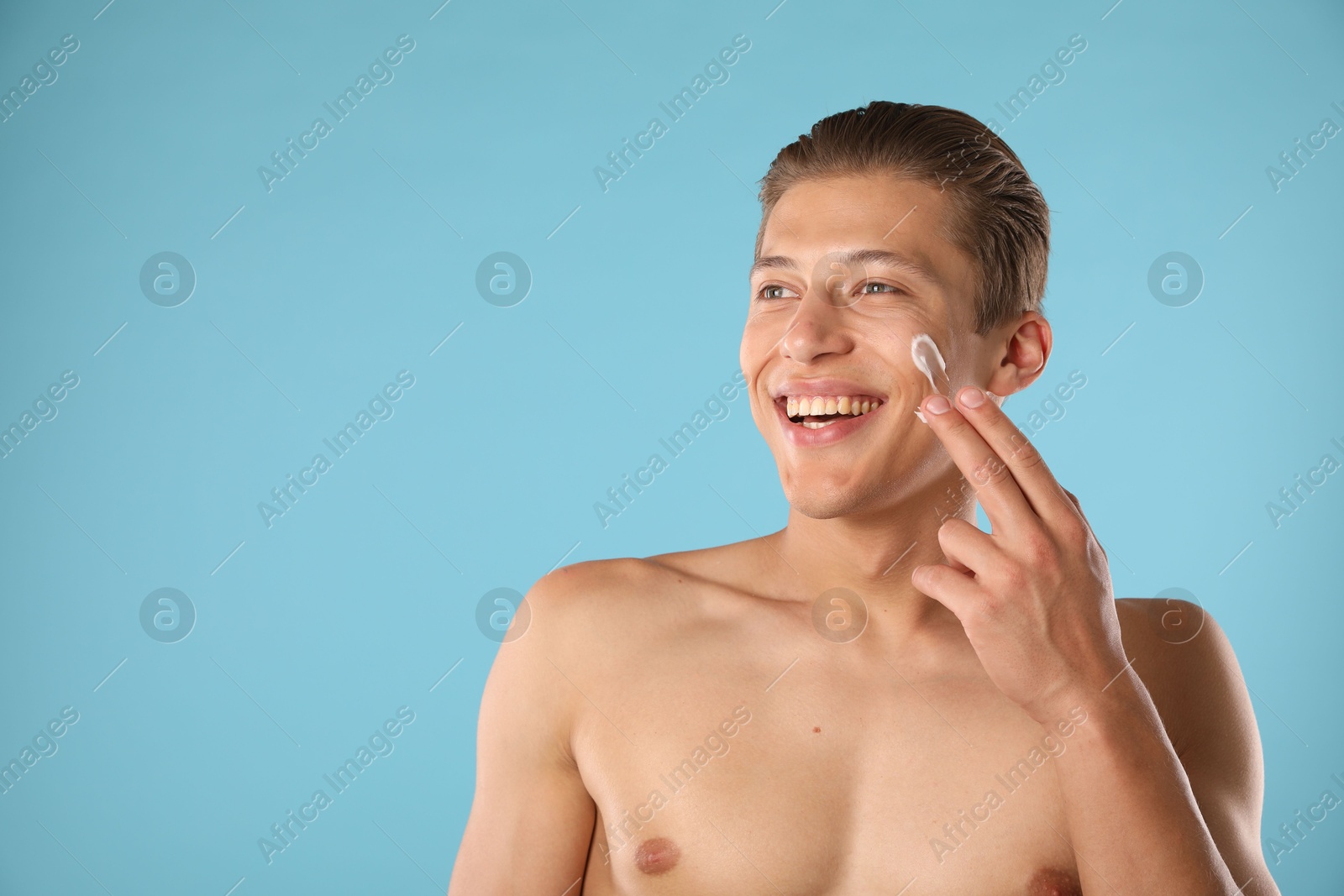 Photo of Handsome young man applying cream onto his face on light blue background, space for text