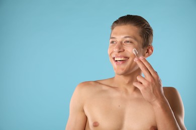 Photo of Handsome young man applying cream onto his face on light blue background, space for text