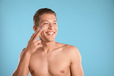 Photo of Handsome young man applying cream onto his face on light blue background, space for text