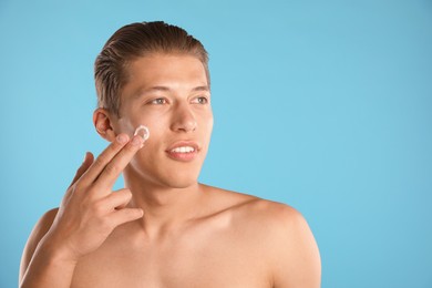 Photo of Handsome young man applying cream onto his face on light blue background, space for text