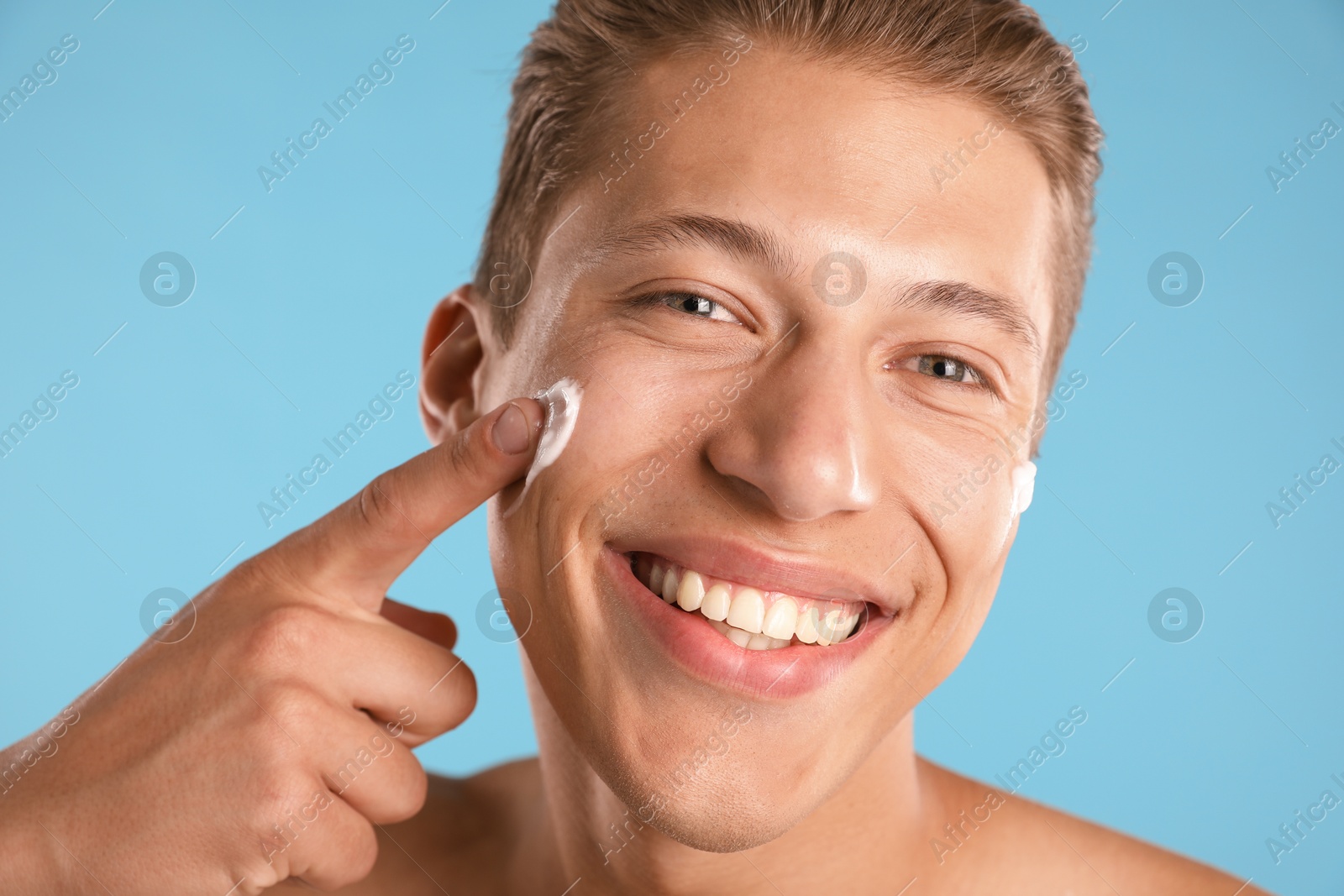 Photo of Handsome young man applying cream onto his face on light blue background, closeup