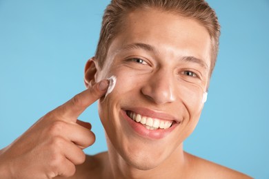 Photo of Handsome young man applying cream onto his face on light blue background, closeup