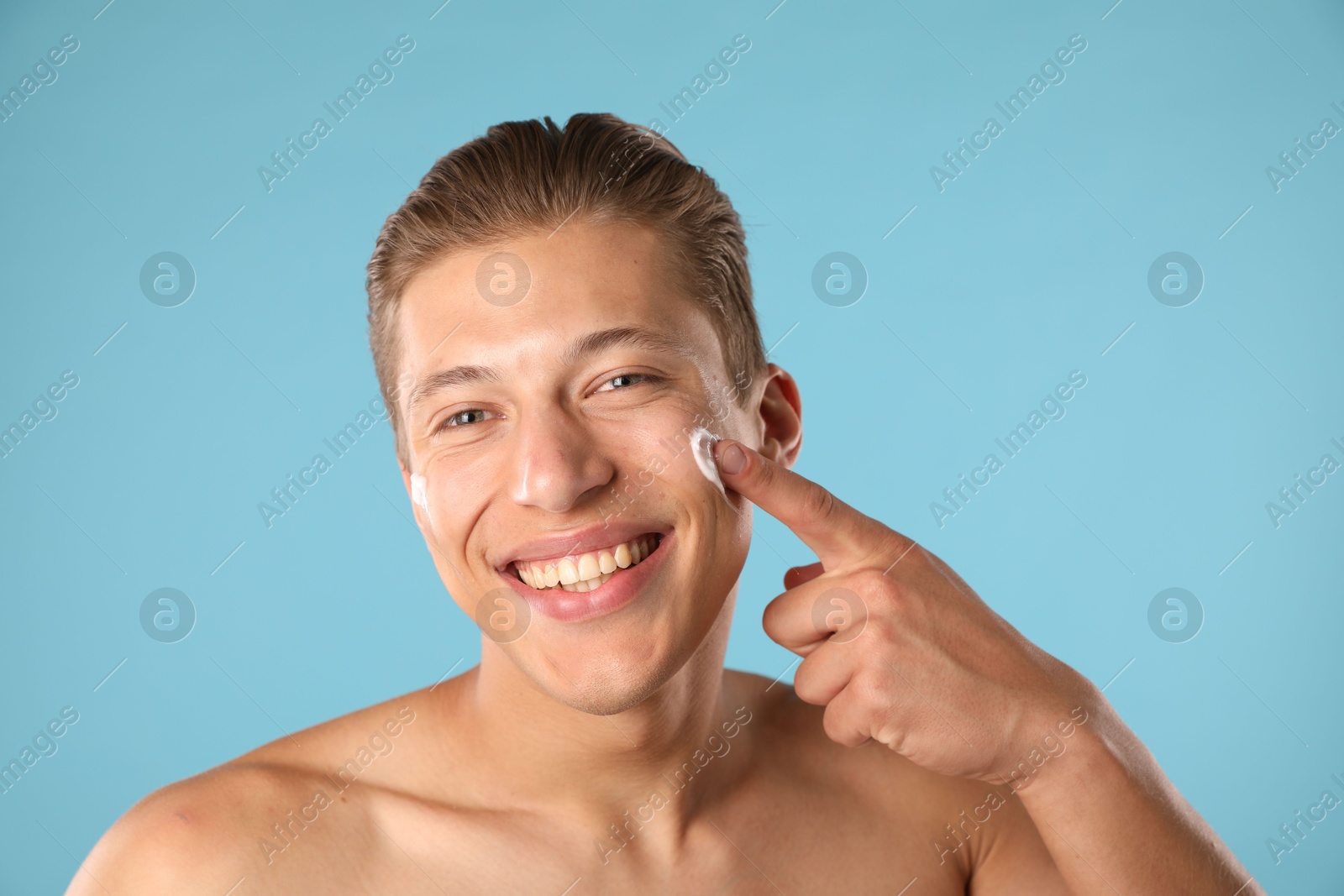 Photo of Handsome young man applying cream onto his face on light blue background, closeup