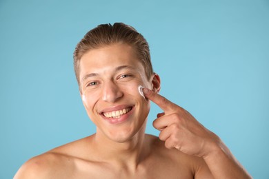 Photo of Handsome young man applying cream onto his face on light blue background, closeup