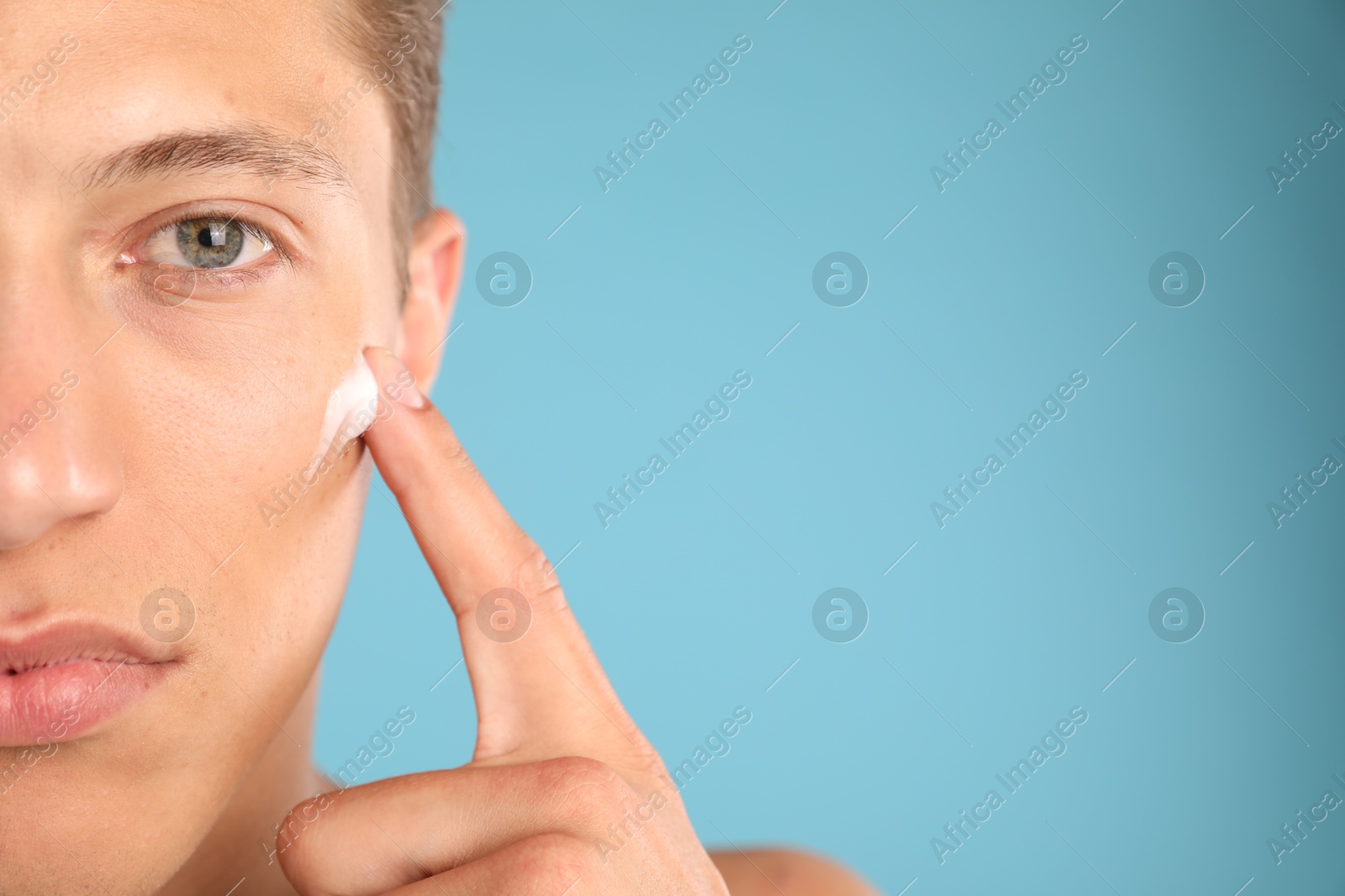 Photo of Handsome young man applying cream onto his face on light blue background, closeup. Space for text