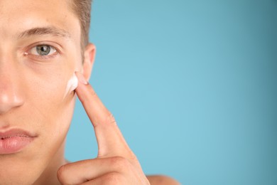 Photo of Handsome young man applying cream onto his face on light blue background, closeup. Space for text